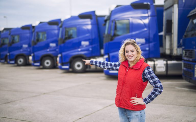 Uśmiechnięta pracownica firmy logistycznej wskazuje ręką na rząd niebieskich samochodów ciężarowych, które wykonują profesjonalny transport farmaceutyczny w Bydgoszczy i regionie.