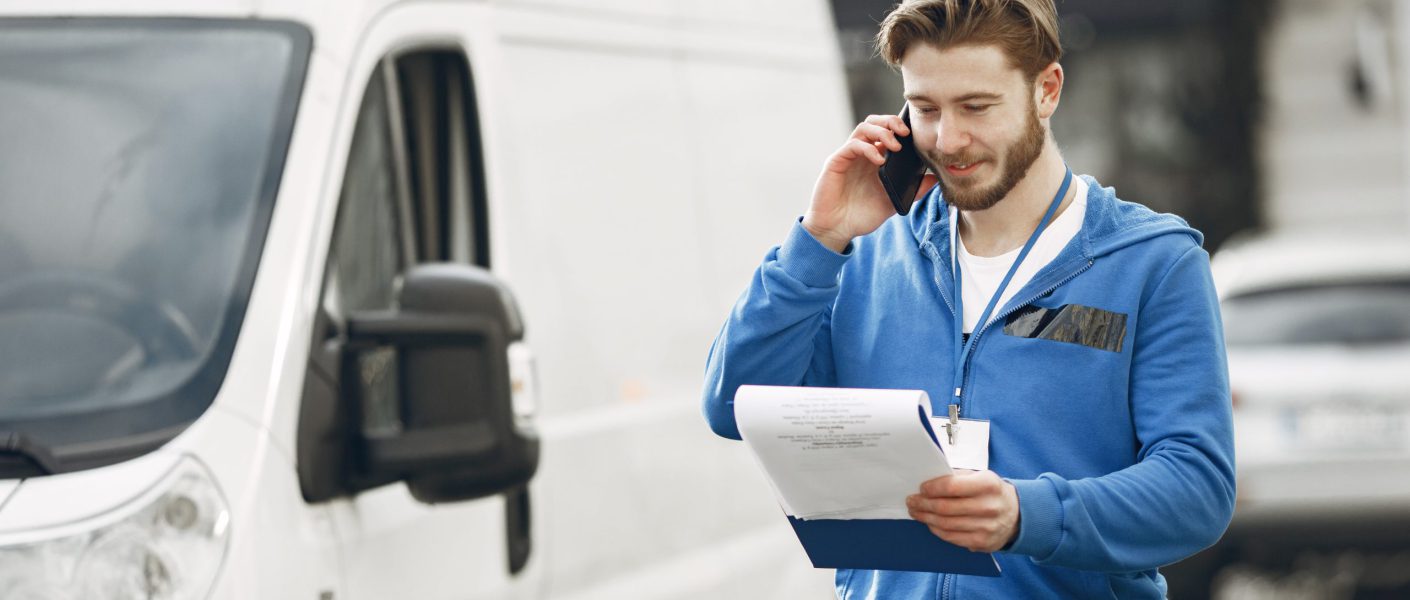 Kierowca w niebieskiej bluzie, współpracujący z firma spedycyjna Bydgoszcz, rozmawia przez telefon i sprawdza dokumenty przewozowe stojąc przy białym samochodzie dostawczym.