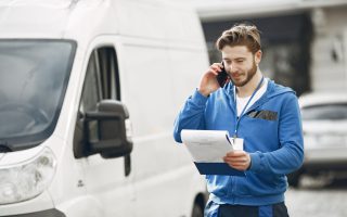 Kierowca w niebieskiej bluzie, współpracujący z firma spedycyjna Bydgoszcz, rozmawia przez telefon i sprawdza dokumenty przewozowe stojąc przy białym samochodzie dostawczym.