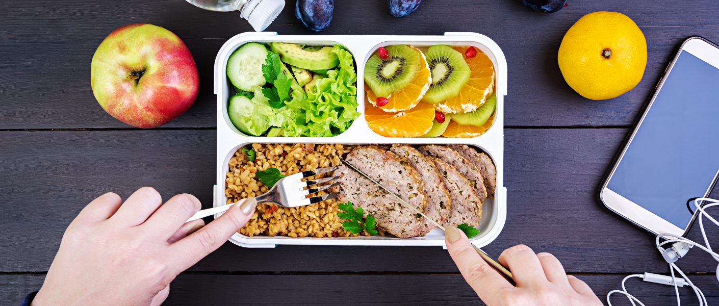 Ujęcie z góry przedstawiające ręce jedzące zdrowy lunch z pudełka umieszczonego na ciemnym, drewnianym stole. W pudełku znajduje się danie główne (mięso i kasza bulgur), świeże warzywa (ogórek, awokado, sałata) oraz owoce (kiwi, pomarańcza), spełniające zasady Dieta pudełkowa bez glutenu i laktozy.