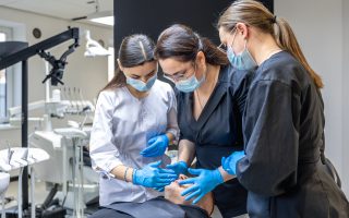 Zespół trzech lekarzy dentystów w maseczkach i rękawiczkach, w tym implanty specjaliści, dokładnie bada jamę ustną pacjenta leżącego na fotelu stomatologicznym w nowoczesnym gabinecie.