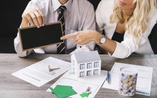 Profesjonalne spotkanie przy biurku z dokumentami, kluczami i modelem domu, podczas którego omawiane są szczegóły umowy, jaką oferuje skup nieruchomości z lokatorami na dany lokal.