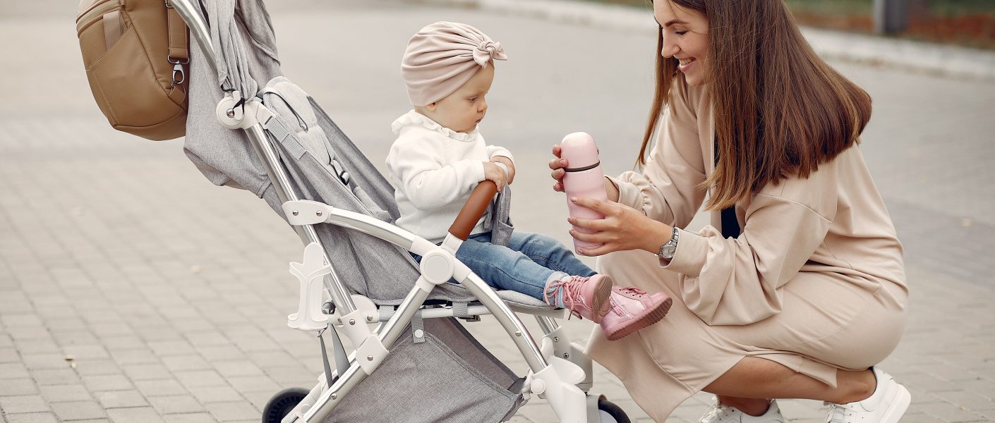 Kobieta kuca przy lekkim wózku spacerowym, podając dziecku termos. Najlepsze sklepy z wózkami Bielsko-Biała oferują nie tylko pojazdy, ale i szeroką gamę akcesoriów ułatwiających jesienne spacery.