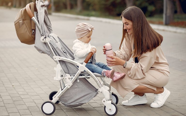 Kobieta kuca przy lekkim wózku spacerowym, podając dziecku termos. Najlepsze sklepy z wózkami Bielsko-Biała oferują nie tylko pojazdy, ale i szeroką gamę akcesoriów ułatwiających jesienne spacery.