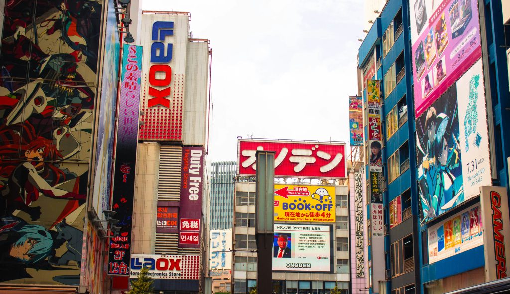 Na zdjęciu widać tętniącą życiem ulicę w japońskiej dzielnicy handlowej, pełną wysokich budynków pokrytych kolorowymi billboardami i reklamami