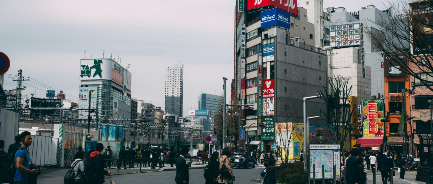 Na zdjęciu widać miejskie skrzyżowanie z przejściem dla pieszych, po którym przechodzą ludzie w codziennych i biznesowych strojach