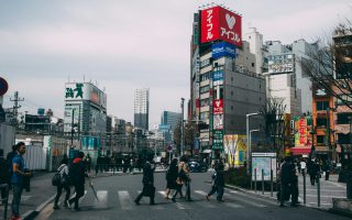 Na zdjęciu widać miejskie skrzyżowanie z przejściem dla pieszych, po którym przechodzą ludzie w codziennych i biznesowych strojach