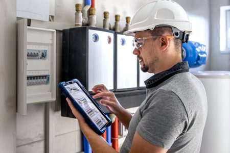 Mężczyzna w kasku i okularach ochronnych obsługuje system instalacji niskoprądowej. Technik trzyma w rękach tablet z widocznym schematem rozdzielni, stojąc przed otwartą skrzynką elektryczną.