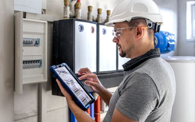 Mężczyzna w kasku i okularach ochronnych obsługuje system instalacji niskoprądowej. Technik trzyma w rękach tablet z widocznym schematem rozdzielni, stojąc przed otwartą skrzynką elektryczną.