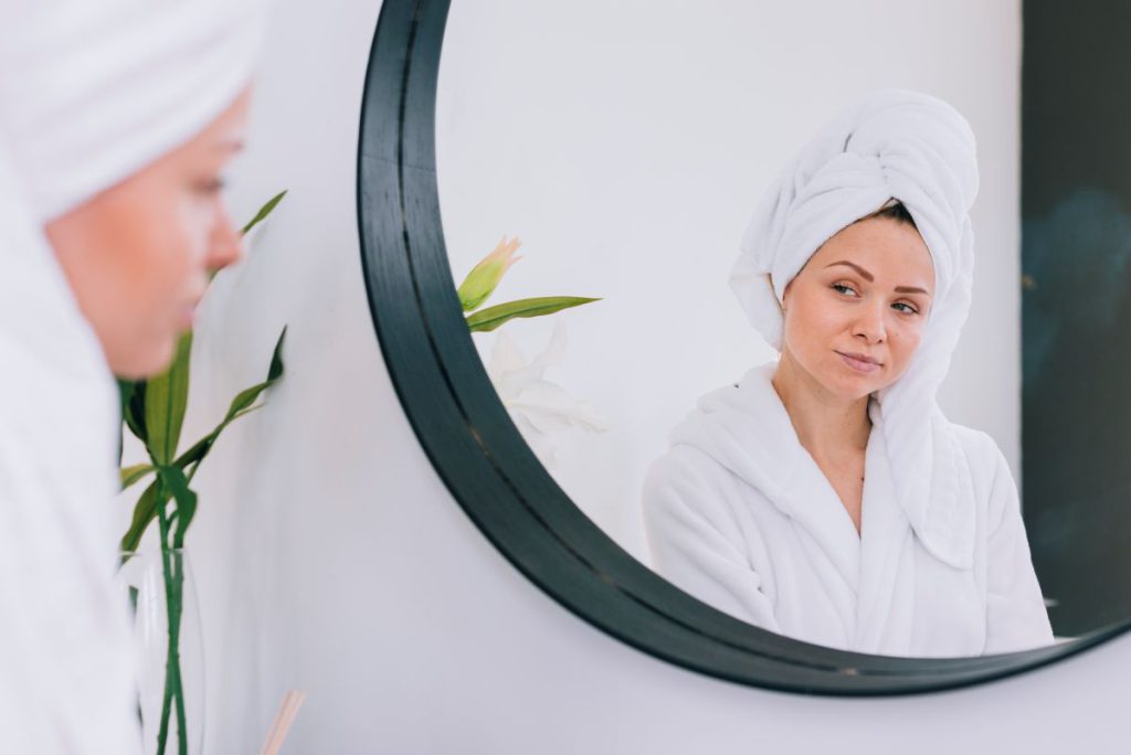 Kobieta w białym szlafroku i ręczniku na głowie przegląda się w dużym, okrągłym lustrze z czarną ramą. Zdjęcie prezentuje lustra na wymiar w Krakowie w minimalistycznej aranżacji domowego SPA.