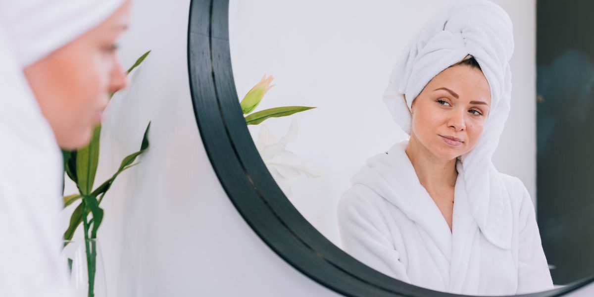 Kobieta w białym szlafroku i ręczniku na głowie przegląda się w dużym, okrągłym lustrze z czarną ramą. Zdjęcie prezentuje lustra na wymiar w Krakowie w minimalistycznej aranżacji domowego SPA.