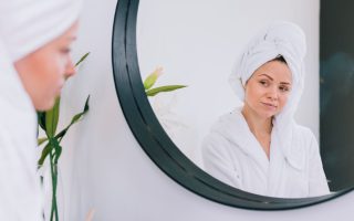 Kobieta w białym szlafroku i ręczniku na głowie przegląda się w dużym, okrągłym lustrze z czarną ramą. Zdjęcie prezentuje lustra na wymiar w Krakowie w minimalistycznej aranżacji domowego SPA.