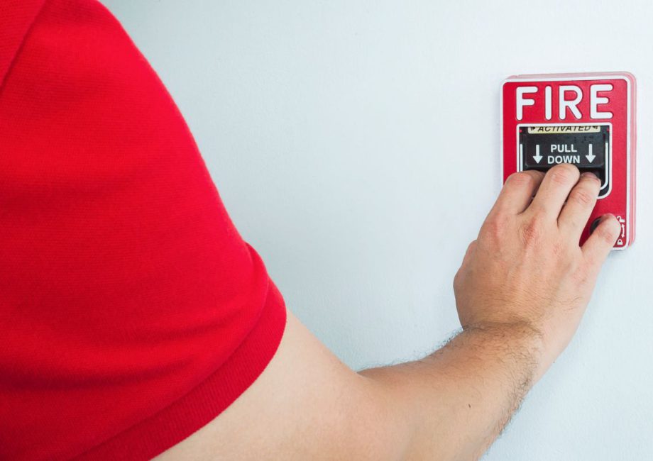 Ręka osoby w czerwonej koszulce pociąga za dźwignię ręcznego ostrzegacza pożarowego na ścianie. Zdjęcie przedstawia testy sprawności urządzeń, jakie przeprowadza serwis instalacji przeciwpożarowych.