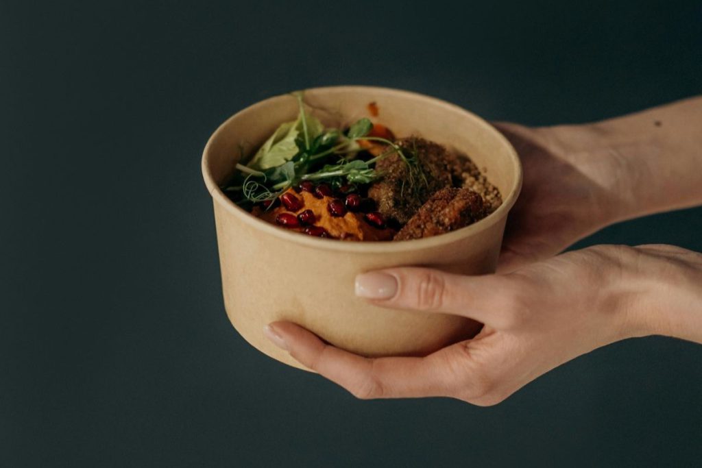 Osoba trzymająca papierową miskę z bogatym w proteiny posiłkiem roślinnym: falafle, hummus z granatem i kasza, które oferuje wysokobiałkowa dieta wegetariańska.