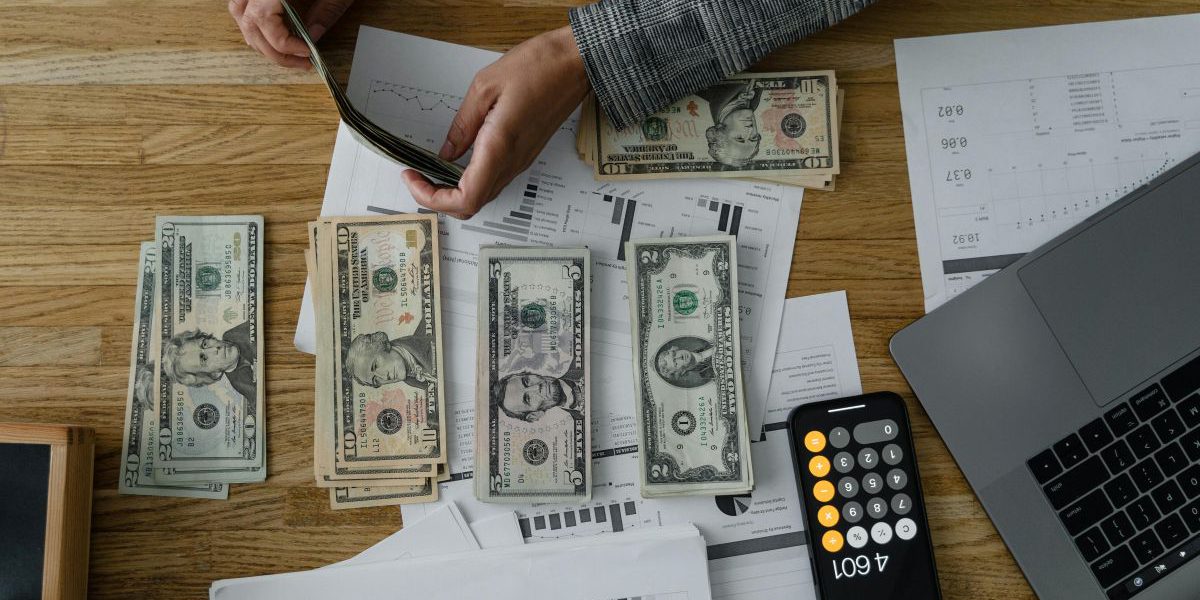 Doradca finansowy Wrocław analizujący wykresy i dokumenty finansowe, przeliczający gotówkę przy użyciu kalkulatora w telefonie i laptopa.