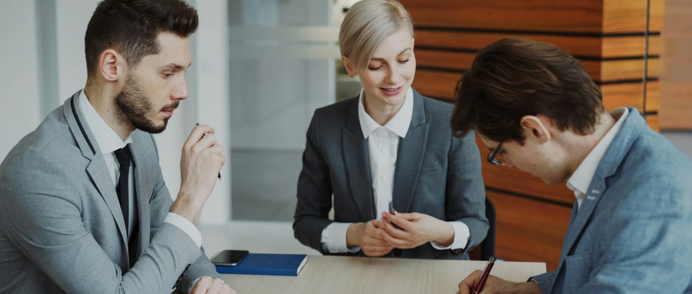 Na zdjęciu przedstawiono troje osób w eleganckich strojach biznesowych podczas podpisywania dokumentu.