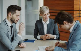 Na zdjęciu przedstawiono troje osób w eleganckich strojach biznesowych podczas podpisywania dokumentu.