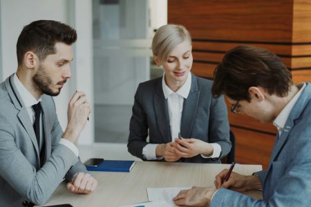 Na zdjęciu przedstawiono troje osób w eleganckich strojach biznesowych podczas podpisywania dokumentu.