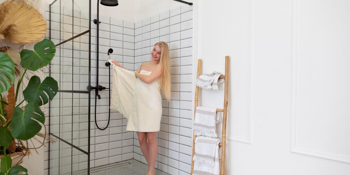 Elegancka łazienka, w której zainstalowano kabiny prysznicowe na wymiar (Kraków). Na zdjęciu widać prysznic typu walk-in z czarnymi profilami i przeszkleniem. W kabinie stoi uśmiechnięta kobieta owinięta białym ręcznikiem.