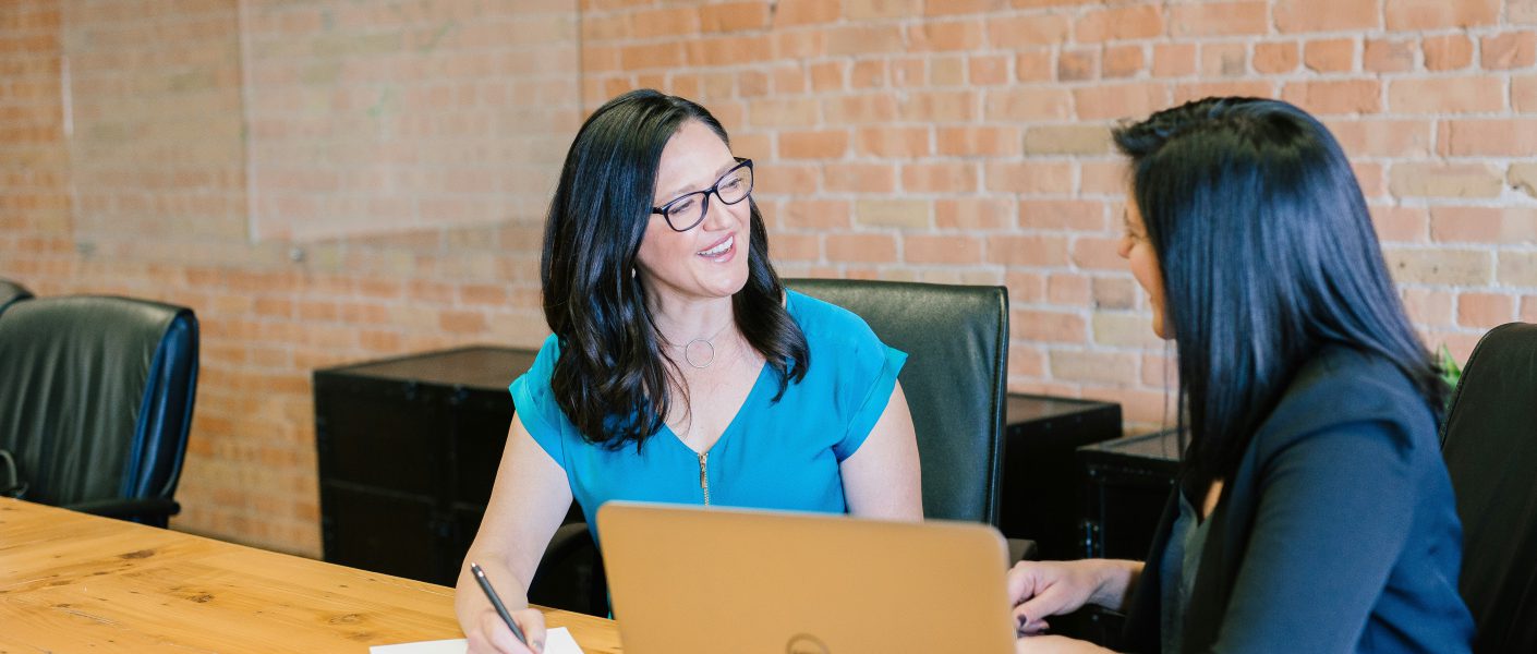 Na zdjęciu widać dwie kobiety siedzące przy drewnianym stole w nowoczesnym biurze z ceglaną ścianą w tle. Jedna z nich notuje coś w notesie, druga siedzi obok z otwartym laptopem